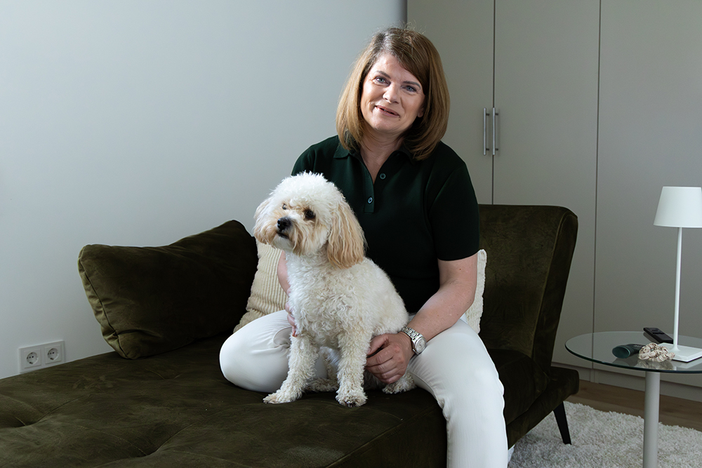 Katja Böcker sitzt mit ihrem weißen Hund auf einem olivgrünen Sofa in einem modernen, hellen Raum