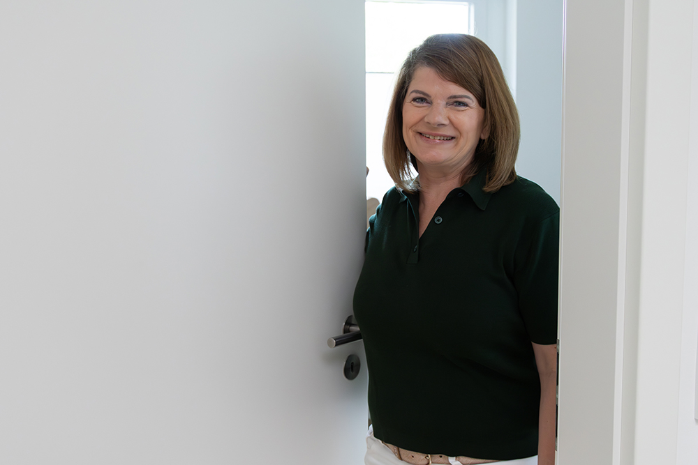 Katja Böcker öffnet lächelnd eine weiße Tür und trägt ein dunkelgrünes Polo-Shirt